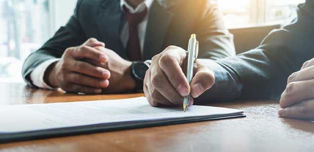 Hand signing a document with someone sitting in background