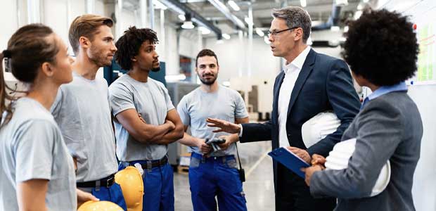 five people watching one person in grey suit talk in industrial plant